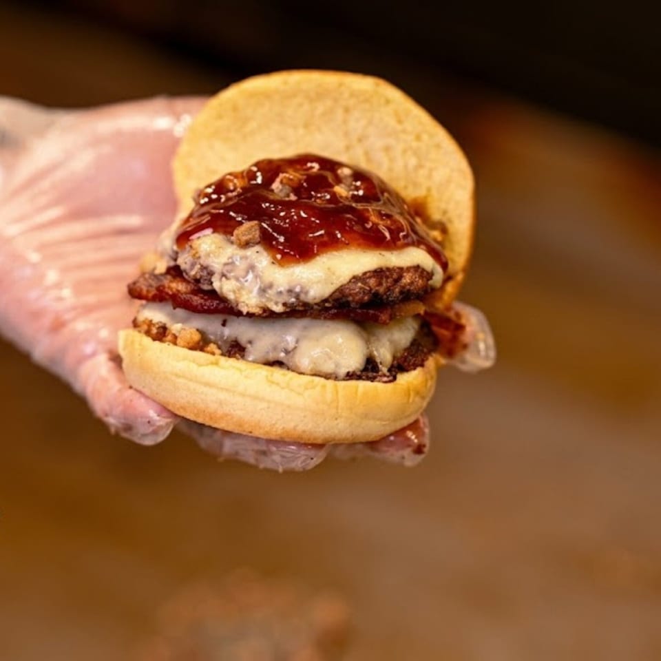 Gloved hand holding a double patty burger with melted cheese, bacon, and BBQ sauce.