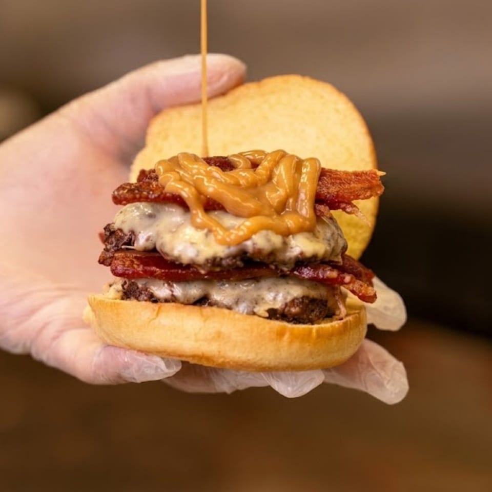 Gloved hand holding a double cheeseburger with bacon and a drizzle of peanut butter sauce.