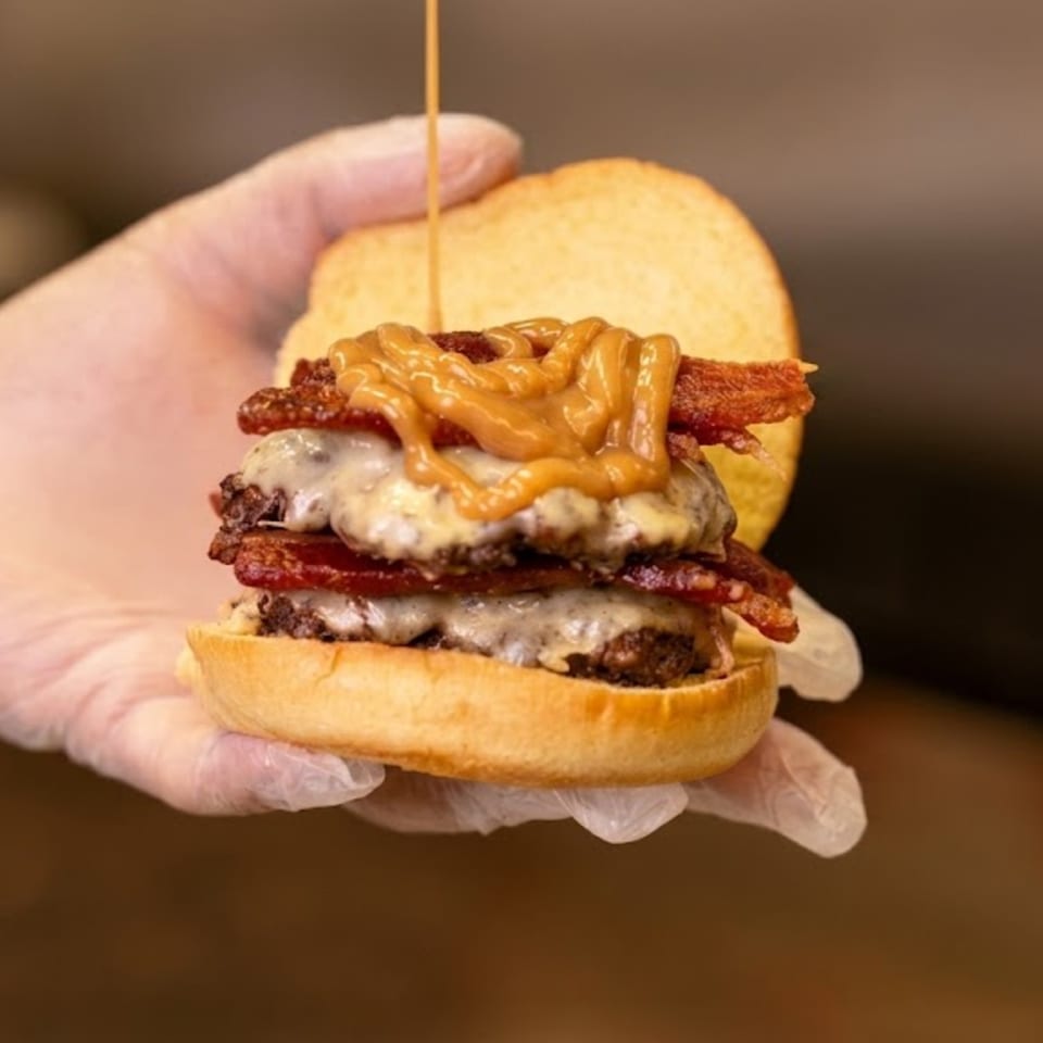 Gloved hand holding a double cheeseburger with bacon and a drizzle of peanut butter sauce.
